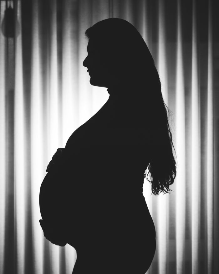 Monochrome silhouette of a pregnant woman standing in profile against curtain backdrop.